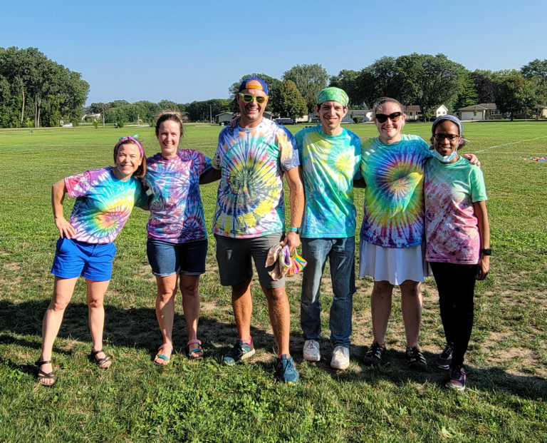 6 fellows and faculty members standing in a row in a green field with a blue sky wearing tie dye shirts