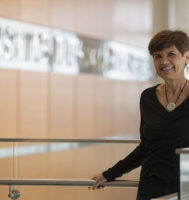 Anita Laxova standing next to a railing in an atrium with light wood paneling walls.