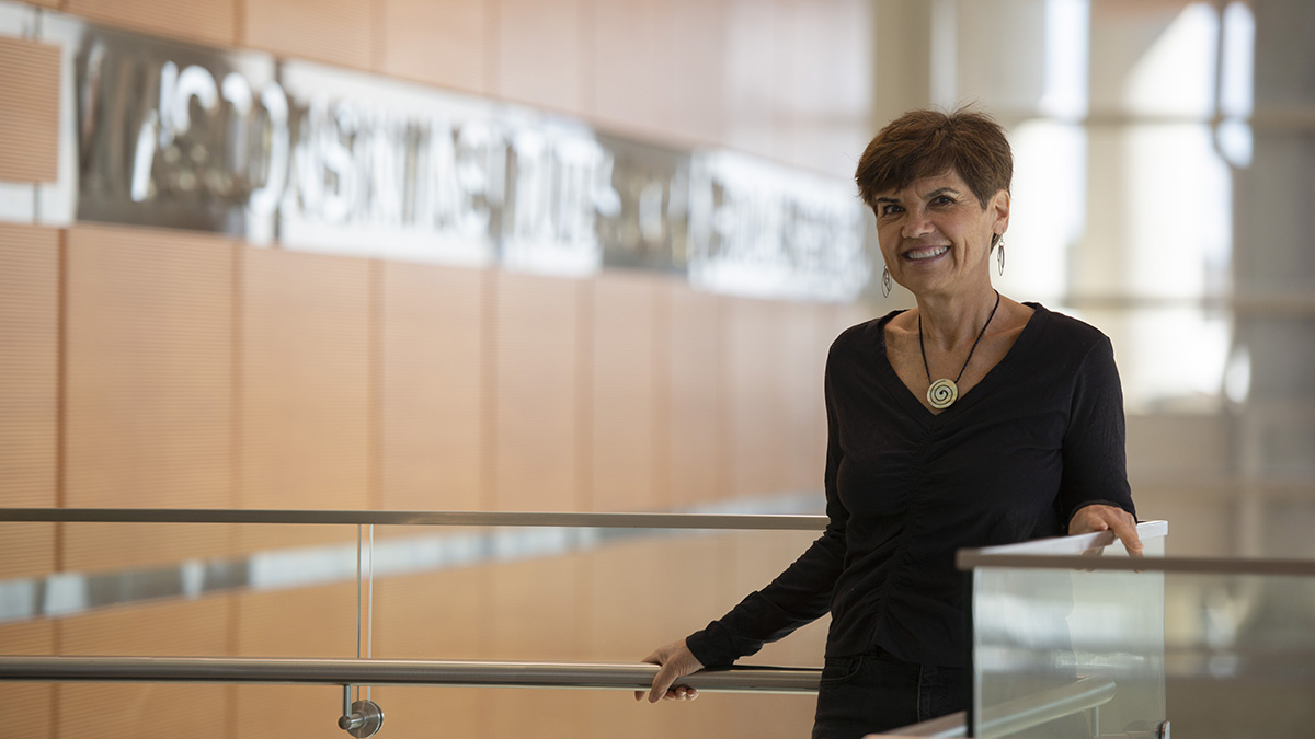 Anita Laxova standing next to a railing in an atrium with light wood paneling walls.