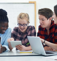 group of teenagers sitting around table looking at laptop computers and mobile devices