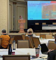 image of person speaking at podium in front of a crown of people sitting at tables