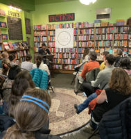 Audience members sitting in folding chairs around the speaker in the front of the room. The walls are filled with books and audience members are attentively listening.