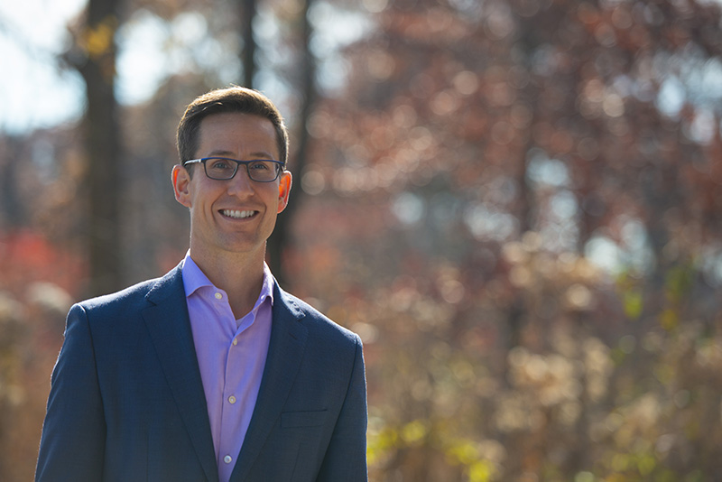 Ryan McAdams smiling at the camera and standing outside with a blurred background of colorful fall trees