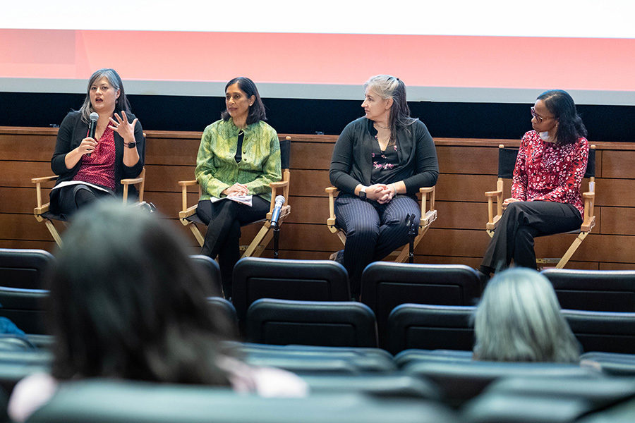 four people sitting in directors chairs on a stage with the person on the far left speaking in to a microphone