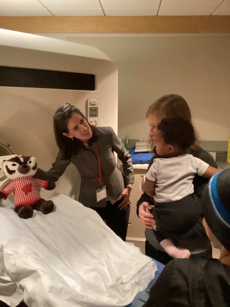 a doctor is showing an MRI machine to a young child being carried by his mother. A stuffed doll of Bucky Badger is on the bed of the machine.