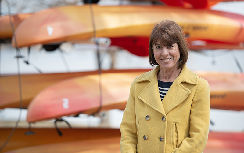 Ann Dodge standing outside in a yellow coat in front of a rack of kayaks