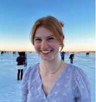 a smiling white woman wearing a lavender top. She is standing on a frozen lake, with several other people and the sunset behind her.