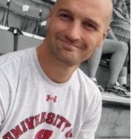 a smiling, bald, white man wearing a University of Wisconsin T-shirt sitting in a stadium