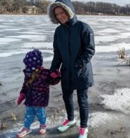a smiling woman holding the hand of a toddler, both in winter coats standing near a frozen pond