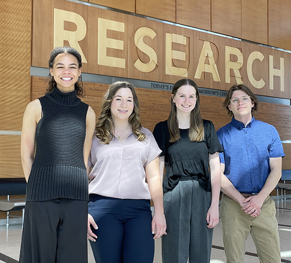 four college-aged students standing under a sign that reads Research