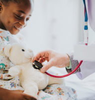 child holding teddy bear while doctor checks the teddy bear's chest with a stethoscope