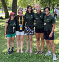 Five young adults wearing matching green camp shirts standing in a row in an outdoors environment.