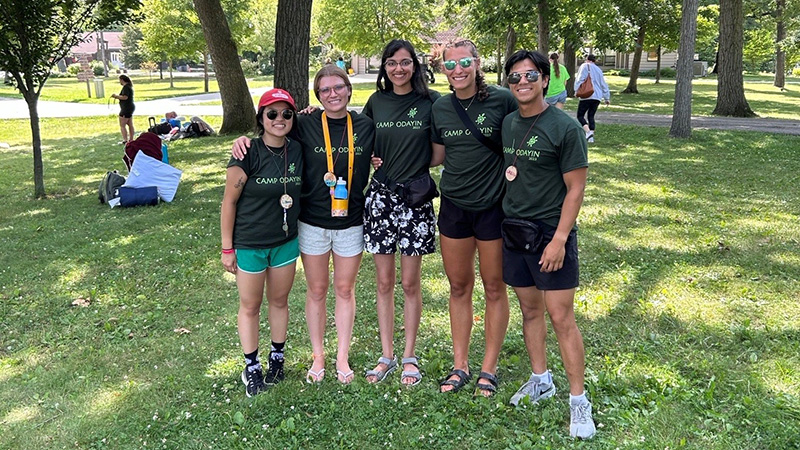 Five young adults wearing matching green camp shirts standing in a row in an outdoors environment.