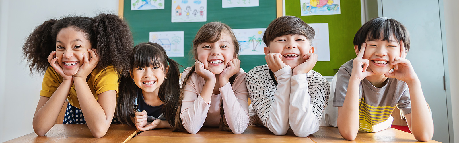 group of five children looking at camera with their chins rested on their hands and their elbows on a table with a chalkboard in the background