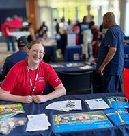 Woman wearing red shirt smiling at camera and sitting at table with pamphlets and small giveaways with a group of people mingling behind her.