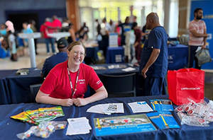 Woman wearing red shirt smiling at camera and sitting at table with pamphlets and small giveaways with a group of people mingling behind her.