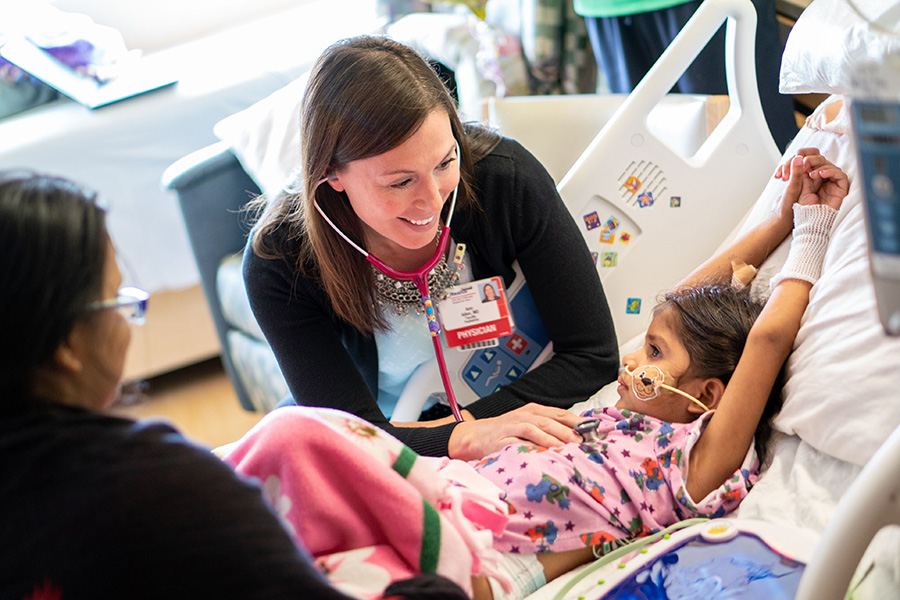 Child in a hospital bed with a doctor leaning over them and smiling while listening to their heart with a stethascope