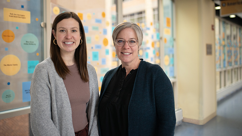 Two health care providers standing in a hallway with windows behind them.