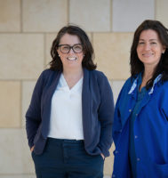 Two doctors standing against a brick wall. One is wearing a pantsuit and the other is wearing scrubs.