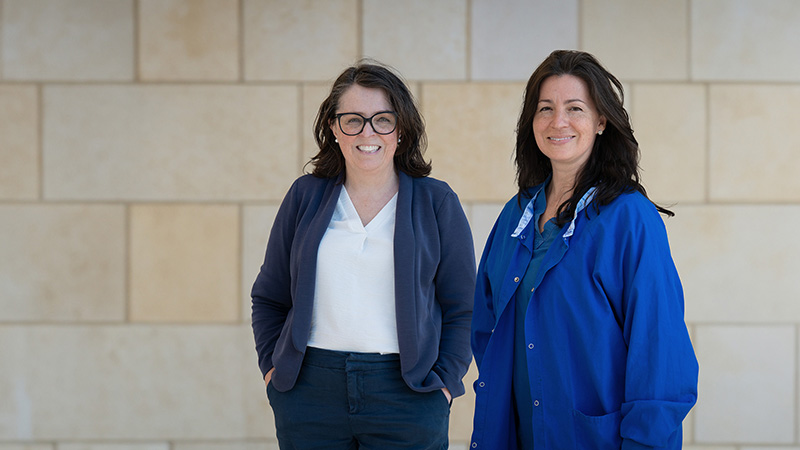 Two doctors standing against a brick wall. One is wearing a pantsuit and the other is wearing scrubs.