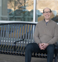 man in a gray shirt sitting on an outside bench and smiling at the camera