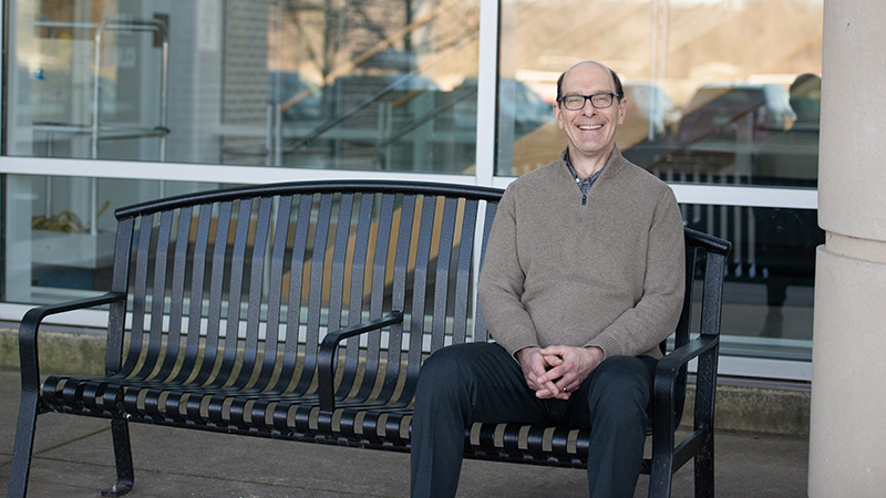 man in a gray shirt sitting on an outside bench and smiling at the camera