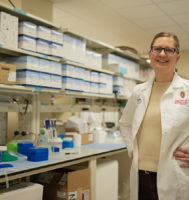Scientist standing in a lab wearing a white lab coat and smiling at the camera