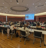dozens of people sitting at desks in a conference room listening to someone speak at the front of the room