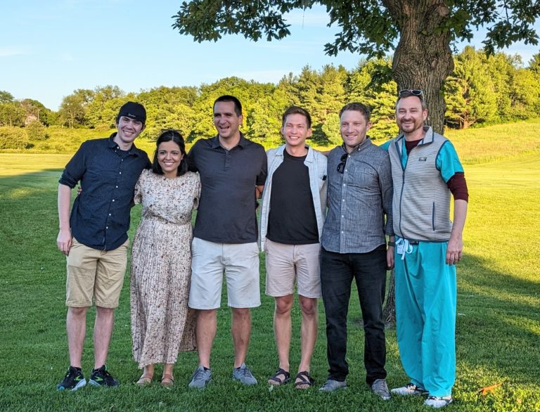 Six fellows standing in a row and smiling in the shade of a tree