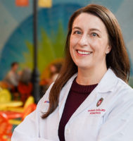 Woman with long brown hair wearing a physician's white coat smiling at camera