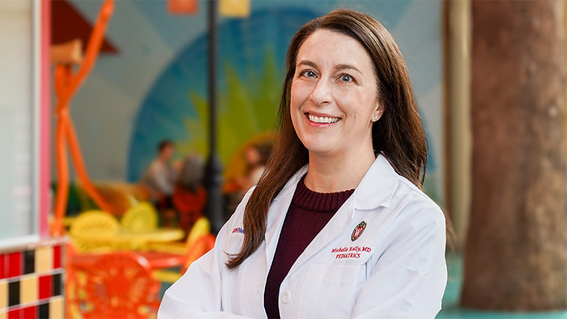 Woman with long brown hair wearing a physician's white coat smiling at camera