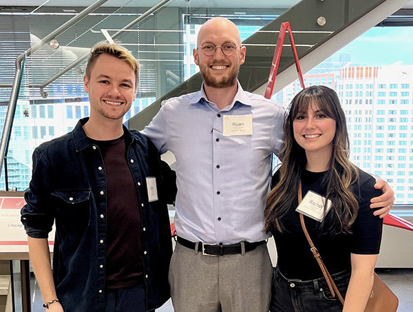 Three people standing in a bright room with a glass staircase behind them.