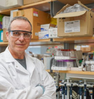 Man standing in a scientific lab wearing a white coat and protective glasses.