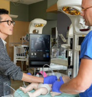 Two doctors standing over a pediatric mannequin on a hospital bed, using and discussing point-of-care ultrasound.