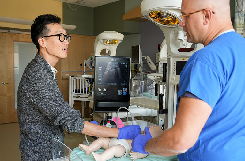 Two doctors standing over a pediatric mannequin on a hospital bed, using and discussing point-of-care ultrasound.