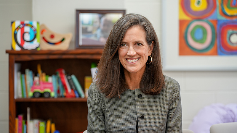 Medical researcher wearing a gray jacket, smiling at the camera, and sitting in a children's clinic area.