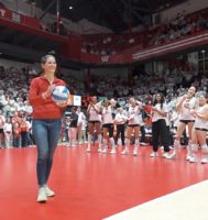 Woman wearing a Badger red shirt and jeans, holding a volleyball and getting ready to serve it across the court.