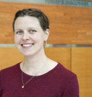 A head-and-shoulders photo of Dr. Tess Woodring standing in an atrium against a faded woodgrain wall. She is wearing a burgundy sweater and smiling at the camera.