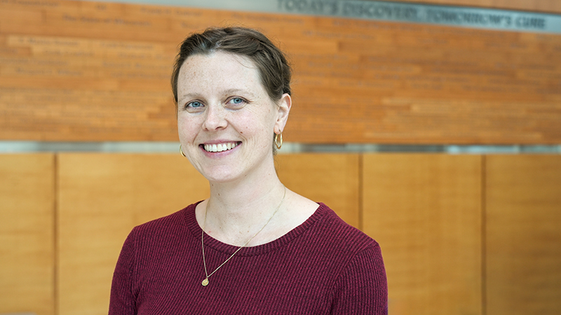 A head-and-shoulders photo of Dr. Tess Woodring standing in an atrium against a faded woodgrain wall. She is wearing a burgundy sweater and smiling at the camera.