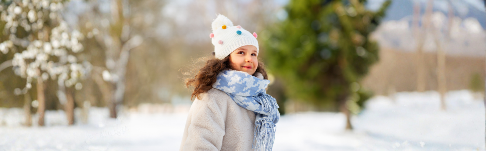 A young girl wearing a white knit hat with colorful accents and a blue scarf stands smiling in a snowy park, with trees softly blurred in the background.