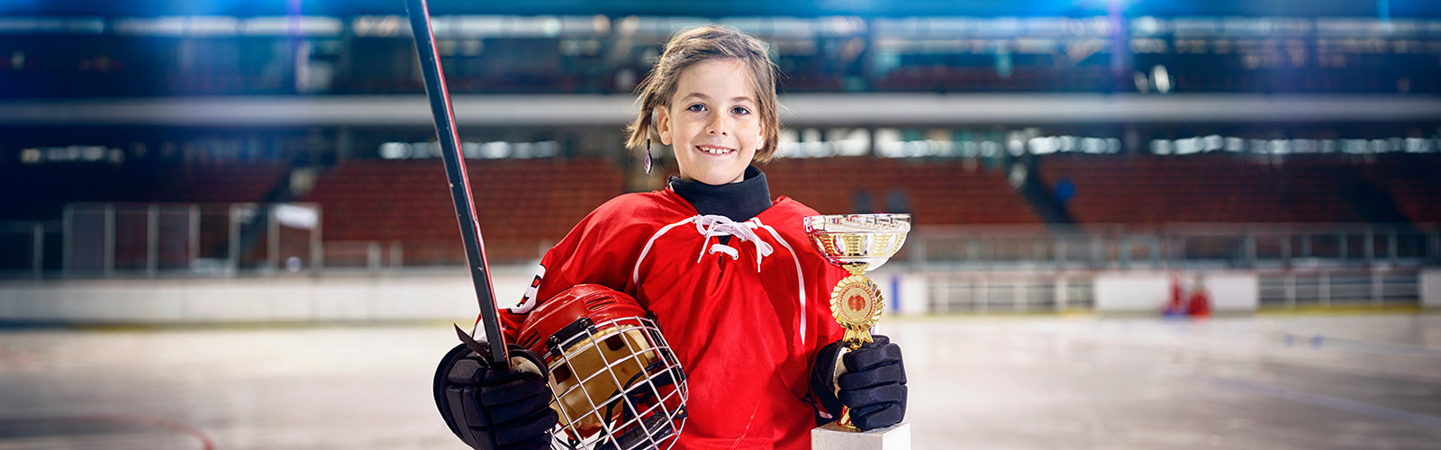 A young hockey player in a red jersey stands on an indoor ice rink, smiling while holding a hockey stick in one hand and a gold trophy in the other, with empty stadium seats in the background.