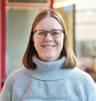 Kim Squires is wearing a light gray turtleneck sweater and red glasses and smiling at the camera.