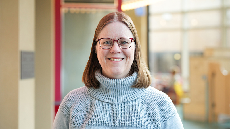 Kim Squires is wearing a light gray turtleneck sweater and red glasses and smiling at the camera.