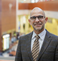 Dr. Norell Rosado is wearing a suite and standing in an atrium while he smiles at the camera.