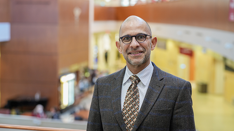 Dr. Norell Rosado is wearing a suite and standing in an atrium while he smiles at the camera.