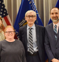Governor Tony Evers stands with three other people against a backdrop of US and Wisconsin state flags.