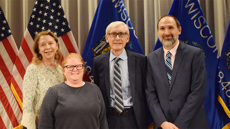 Governor Tony Evers stands with three other people against a backdrop of US and Wisconsin state flags.