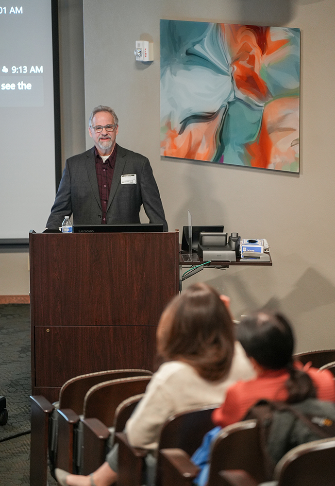 Man with gray hair and beard in a dark suit jacket standing at a podium and talking to an audience.