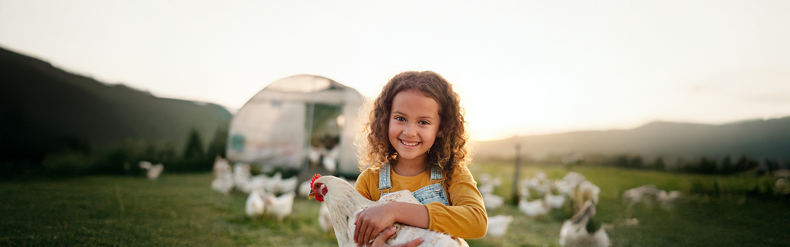Girl with curly hair and overall standing in field holding chicken while smiling.