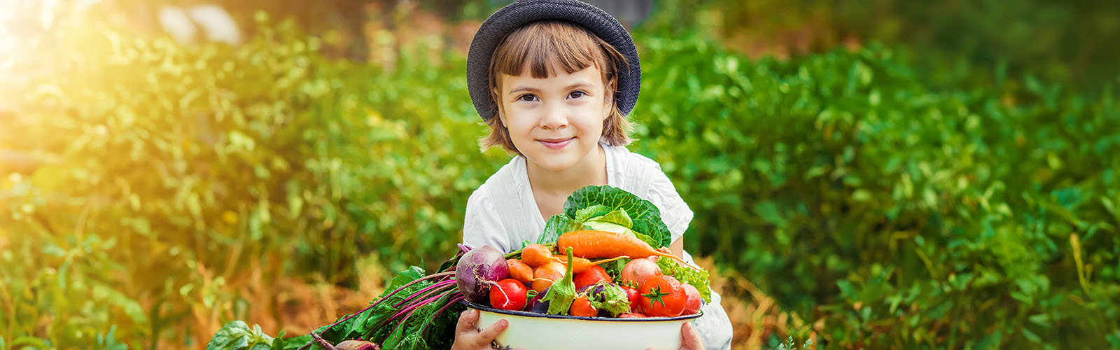 A child wearing a hat is standing in a garden holding a bowl of vegetables.
